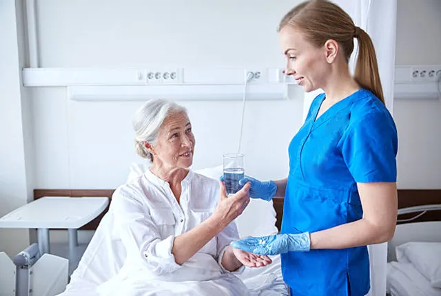 Pflegefachkraft reicht einer Patientin Medikamente und ein Glas Wasser